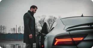 Forretningsmand i mørkegrå frakke står ved den åbne dør til en sort moderne bil lejet på erhvervsleasing.