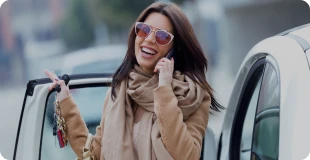 Kvinde med et strålende smil, iført solbriller og et beige halstørklæde, står ved siden af sin nye leasingbil og taler begejstret i telefonen.